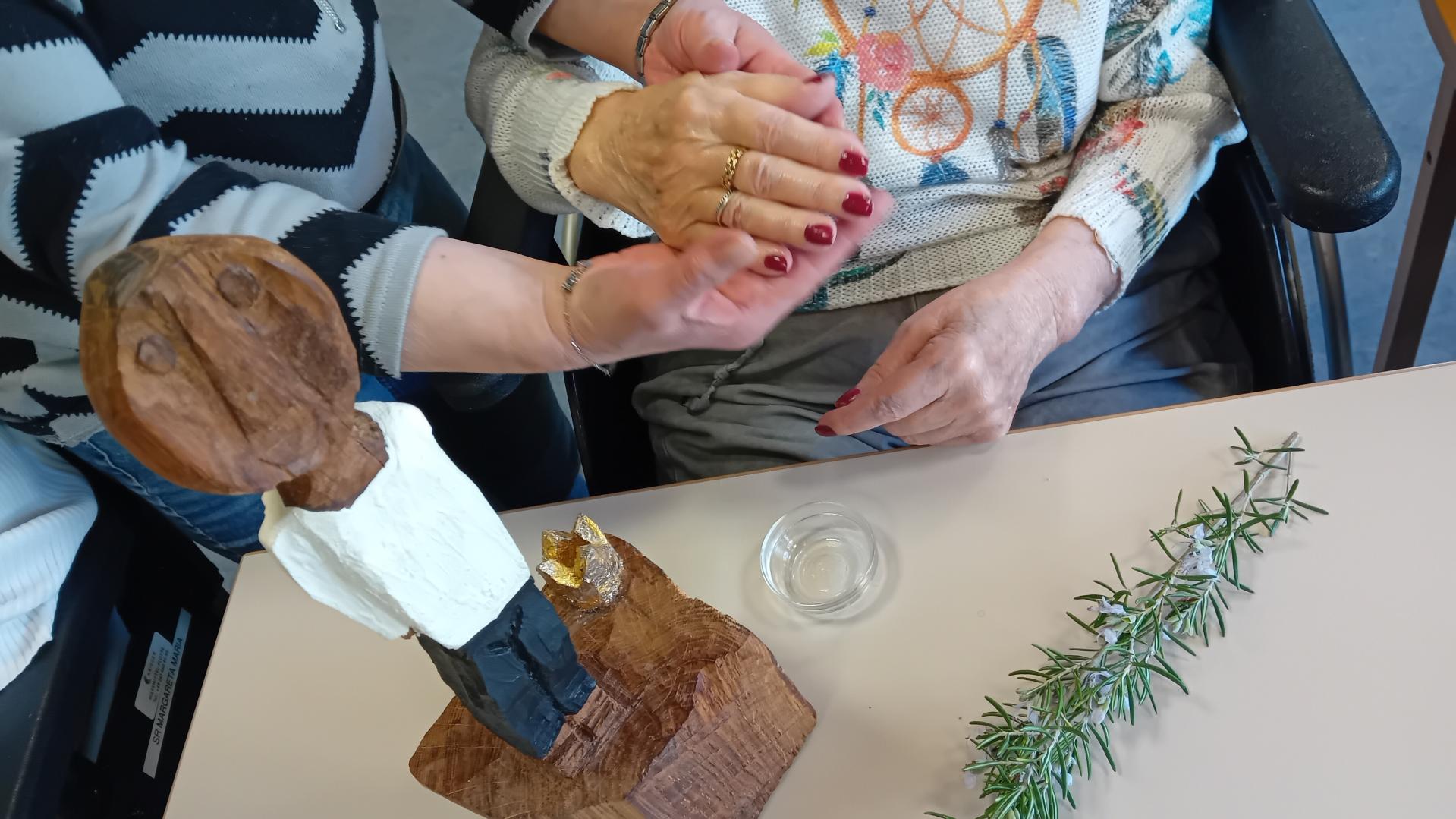 Die kleine Königsfigur steht auf einem Tisch. Daneben werden alte Hände von jüngeren gesalbt.
