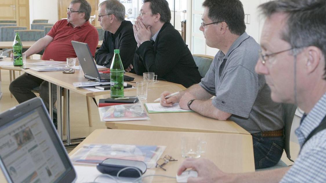 Fünf Männer sitzen - zum Teil mit Laptops - konzentriert an Beratungstischen, die kreisförmig aufgestellt sind.