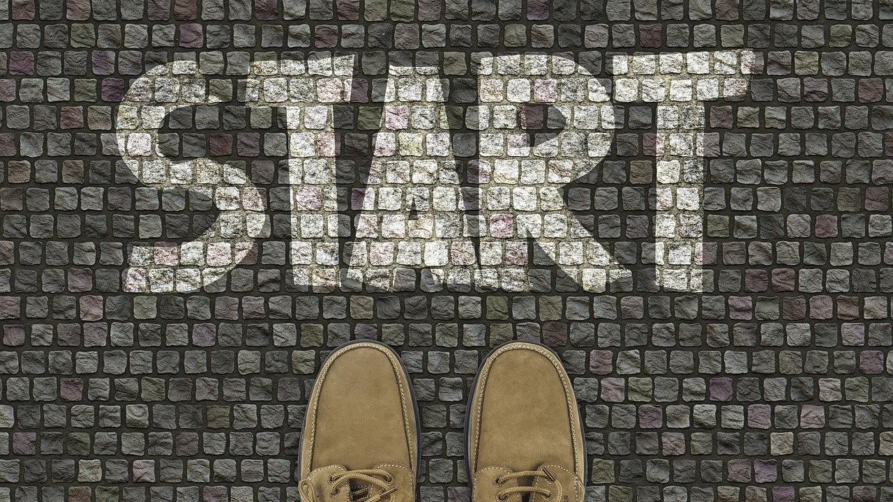 Auf kleinen Pflastersteinen ist mit Kreide das Wort 'Start' geschrieben, Fußspitzen stehen darunter.